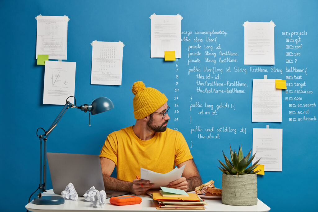 inexperienced european man looks through paper documents, analyzes during working process, wears yellow hat and t shirt, sits at desktop against blue background with papers and written information